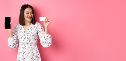 Happy young asian woman internet shopping, showing smartphone screen and looking satisfied at credit card, standing over pink background photo