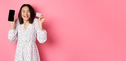 Joyful korean woman showing smartphone screen and credit card, paying for internet order, demonstrating online shopping app, standing over pink background photo