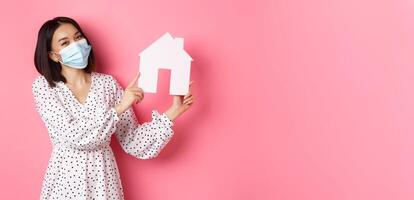 Covid-19, real estate and lifestyle concept. Cute asian woman in face mask selling houses, showing model of home and looking at camera, standing over pink background photo