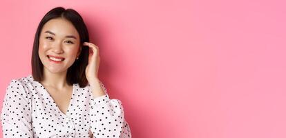 Close-up of beautiful asian woman smiling happy, touching new haircut, standing over pink background photo