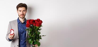 Handsome man in suit going to make a proposal, standing with red roses flowers and engagement rin in box, making romantic surprise, white background photo