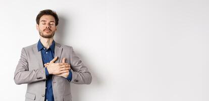 Young man in suit close eyes, holding hands on heart and remember happy memories, feeling nostalgic, standing against white background photo