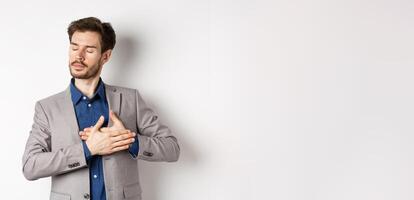 Romantic bearded man in stylish suit holding hands on heart, close eyes and remember something with tender feelings, standing nostalgic on white background photo