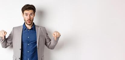 Wow look down. Excited business guy in suit pointing fingers bottom advertisement and look amazed, standing on white background photo