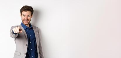 Its you. Smiling handsome manager in suit pointing at camera, choosing person and looking pleased, standing on white background photo