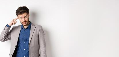 Annoyed male employer scolding personal, pointing finger at head, are you stupid out of your mind gesture, stare at camera from under forehead, standing in suit on white background photo