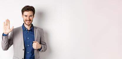 Smiling handsome businessman in suit waving hand, greeting friend with friendly face, say hellp, standing on white background photo