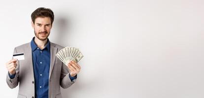 Rich smiling man in suit showing money and plastic credit card, standing with dollar bills and look pleased, white background photo