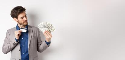 Successful man showing plastic credit card and looking at dollar bills fan, making money deposit, standing on white background photo