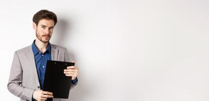 Young male ceo manager holding clipboard with documents and looking at camera, inspecting office, standing on white background photo