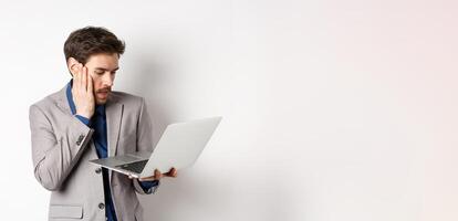 Shocked and worried businessman making mistake on laptop, looking at computer upset, standing on white background photo