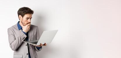Shocked office worker in suit looking worried at laptop screen, having trouble at work, standing on white background photo