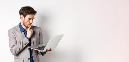 Pensive businessman working on laptop and thinking, looking thoughtful at computer screen, standing in suit on white background photo