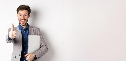 Good job. Happy smiling business man in suit holding laptop, showing thumb up to praise you, nice work gesture, standing on white background photo