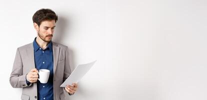 Boss reading documents and drinking morning coffee from mug, looking at paper serious, standing in suit on white background photo