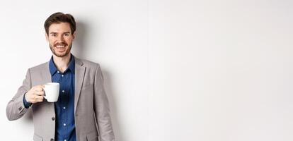 Handsome businessman in suit drinking coffee or tea from office mug, smiling enthusiastic at camera, standing against white background photo