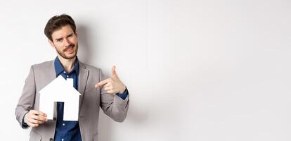 Real estate and insurance concept. Salesman in grey suit showing paper house cutout, selling property, smiling at camera, white background photo