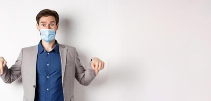 Covid-19, pandemic and business concept. Surprised male ceo manager in medical mask and suit pointing fingers down, looking amused, white background photo