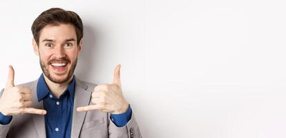 Excited salesman smiling, showing phone signs and asking to give him call, giving his number, standing on white background in suit photo
