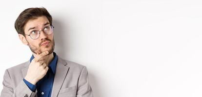 Thoughtful businessman with beard and glasses making decision, looking up and thinking, standing in suit with pensive face, white background photo