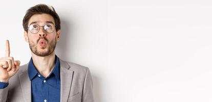 Excited handsome man in glasses and business suit, look up and pointing at top advertisement, exclaim wow with amazed expression, white background photo