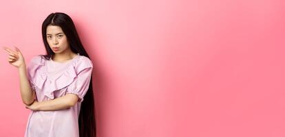 Sad and grumpy asian girl sulking, pointing left and look at camera jealous, want something, complaining and showing desired thing, standing on pink background photo