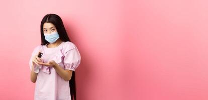 Covid-19, lifestyle and health concept. Beautiful asian woman in medical mask clean hands with sanitizer, apply antiseptic on palms, standing on pink background photo
