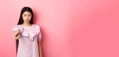 She says no. Serious asian girl shaking finger in stop gesture, prohibit and disagree with person, scolding bad behaviour, standing against pink background photo