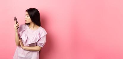 Asian girl look surprised at smartphone screen, reading message, standing in dress against pink background photo