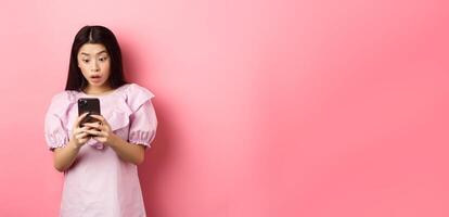 Online shopping. Asian teen girl look surprised at mobile phone screen, reading message with excited expression, standing against pink background photo