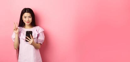 Online shopping. Stylish asian teen girl using smartphone, show peace sign and smiling pleased at camera, standing on pink background photo