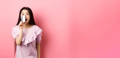 Excited asian girl showing plastic credit card and smiling, going on shopping, standing against pink background photo