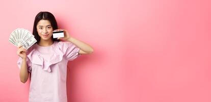 Smiling beautiful asian girl showing money and plastic credit card, giving choice, standing against pink background photo