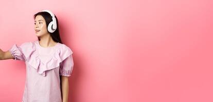 Beautiful asian girl blogger taking selfie in wireless headphones, make photo for social media on smartphone, standing in dress against pink background