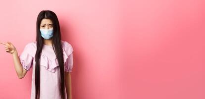 Covid-19, pandemic lifestyle concept. Sad and gloomy asian girl in medical mask pointing left, complain on unfair situation, standing disappointed against pink background photo