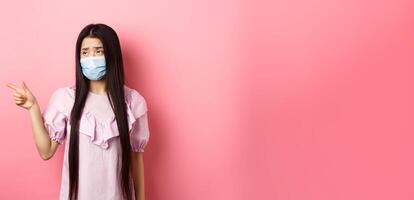 Coronavirus, quarantine and lifestyle concept. Sad and gloomy asian woman in medical mask pointing, looking left upset, showing unfair thing, standing on pink background photo
