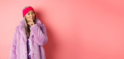 Fashion and shopping concept. Modern chinese senior woman shushing, telling a secret and looking cunning at camera, standing in trendy fake fur coat and headband on pink background photo