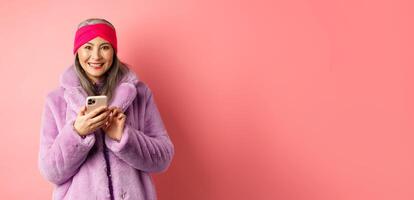 Online shopping and fashion concept. Smiling asian senior lady in winter coat and headband order in internet, holding mobile phone and looking happy, pink background photo