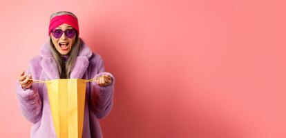 Shopping and fashion concept. Stylish asian elderly woman in sunglasses and faux fur coat open paper bag with gifts, looking amazed at camera, pink background photo