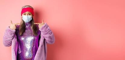 Covid-19, social distancing and fashion concept. Asian edlerly woman in stylish party outfit and respirator, pointing fingers at face mask, asking to follow coronavirus quarantine rules photo