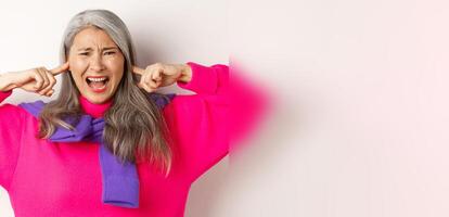 Close-up of annoyed and bothered asian senior woman screaming with shut ears, disturbed by loud noise, standing over white background photo
