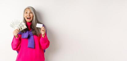 Shopping concept. Happy asian old woman with grey hair, looking fascinated at upper left corner, showing plastic credit card and money dollars, white background photo