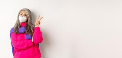 Covid, pandemic and social distancing concept. Cheerful and stylish asian senior woman wearing respirator from coronavirus, showing victory sign, standing over white background photo