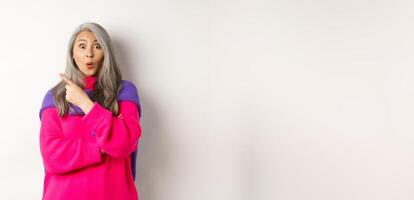 Portrait of impressed asian lady checking out promo offer, pointing at upper left corner and staring at camera amazed, standing in trendy pink sweater over white background photo