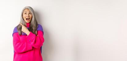Portrait of amazed asian senior woman gasping fascinated, pointing at upper left corner, demonstrate special promotion, standing over white background photo