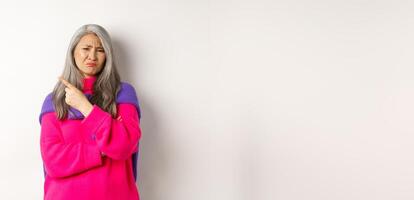 Disappointed asian grandmother grimacing disgusted, pointing finger left and complaining on something bad, standing over white background photo
