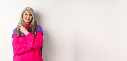 Disappointed asian grandmother grimacing disgusted, pointing finger left and complaining on something bad, standing over white background photo