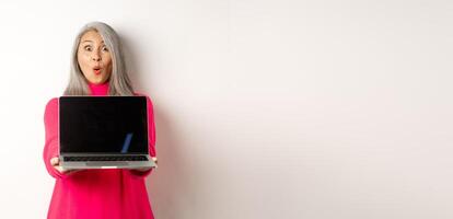 E-commerce concept. Impressed asian senior woman showing blank laptop screen and staring amazed at camera, standing over white background photo