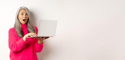 Surprised and impressed asian grandmother staring shocked at camera after reading news on laptop screen, standing over white background photo