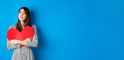 Valentines day. Sad heartbroken girl sobbing, looking aside at empty space and crying upset, hugging big red heart, feeling lonely without lover, standing alone on blue background photo